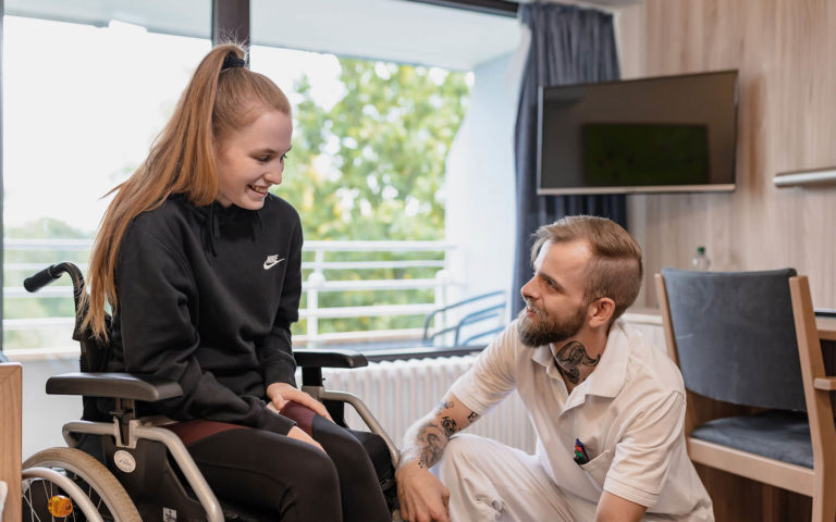 Patientin sitzt im Rollstuhl und Pfleger spricht freundlich mit ihr